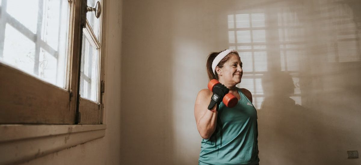 Senior woman in a brightly lit room exercises with dumbbells, promoting fitness at home.